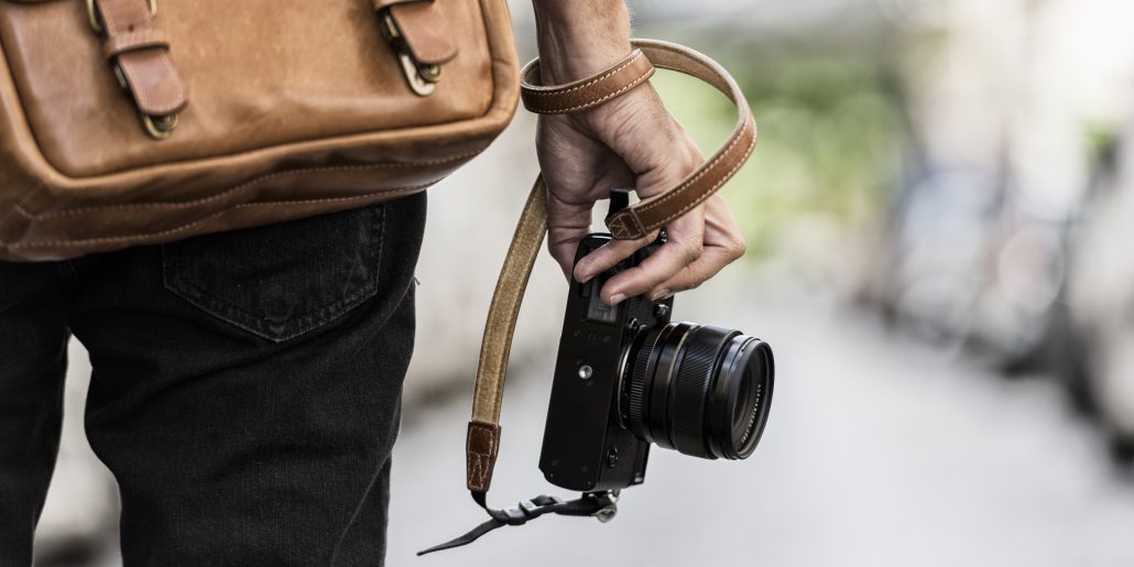 Photographer wearing a brown bag holding a black camera with straps warped around his wrist. 